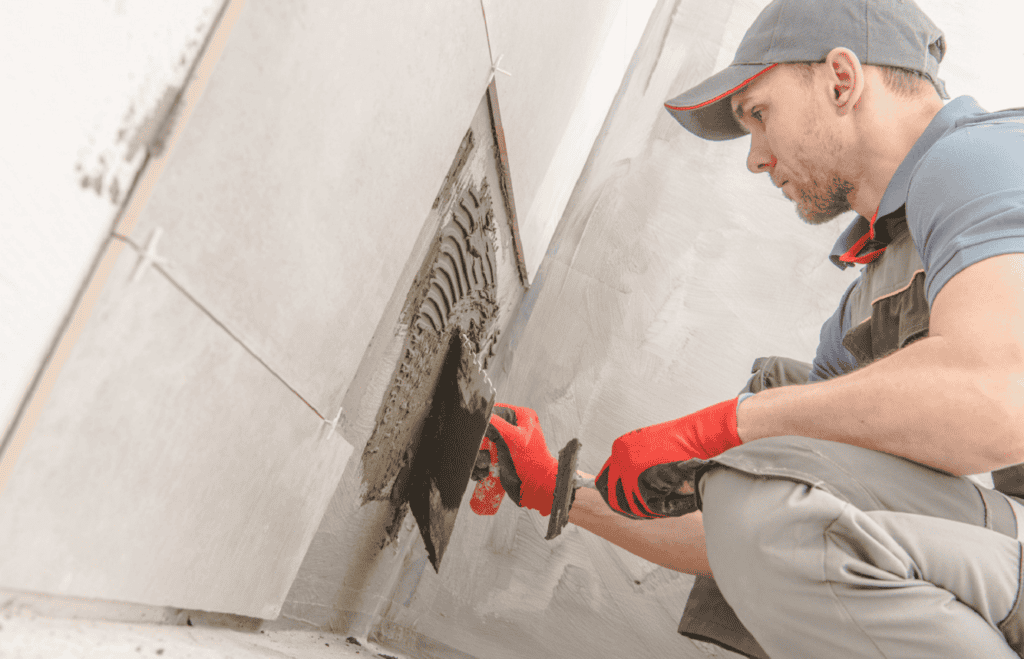 A construction worker in a gray cap and red gloves applies mortar to a section of a tiled wall using a trowel. Specializing in bathroom remodeling Orlando Florida, the worker carefully fills gaps between the partially installed ceramic tiles, ensuring precision and quality in every stroke.