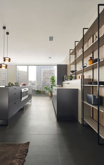 A modern kitchen with minimalistic design, featuring sleek dark grey cabinets and a large island, newly crafted by Kitchen Remodeling Orlando Florida. Shelving is stocked with various kitchen items. Large floor-to-ceiling windows let in natural light, offering a view of city buildings outside.