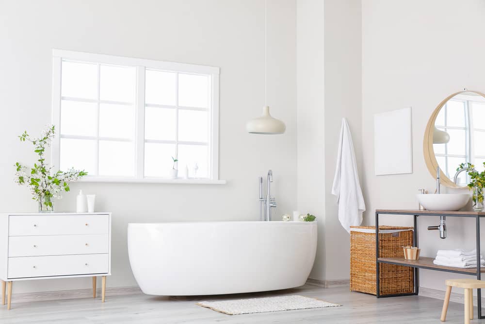 Modern minimalist bathroom with a freestanding white bathtub in front of a large window. A pendant light hangs above. To the right, a wicker hamper and a round mirror above sleek bathroom countertops Orlando Florida residents adore. A potted plant and small dresser add fresh greenery.