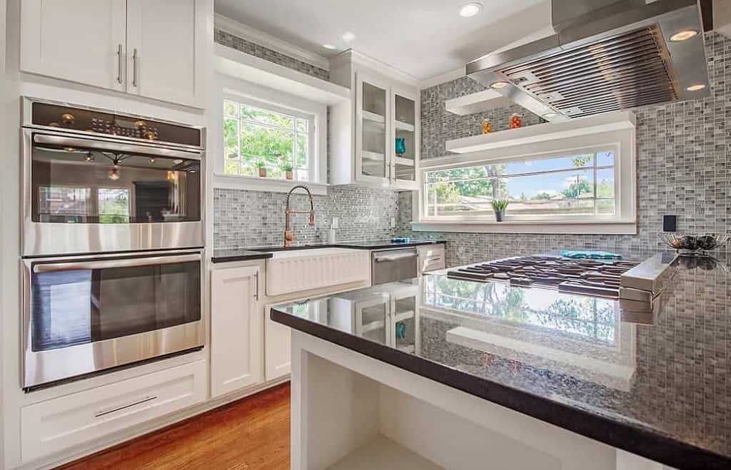 A modern kitchen in Orlando, Florida, features white cabinets, a stainless steel double oven, and a large gas stovetop on a black granite island. The gray mosaic tile backsplash complements the farmhouse sink, while open shelves and recessed lighting add to the sleek kitchen design by General Contractor Orlando Florida.