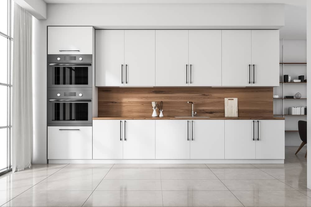 A modern kitchen with white cabinetry, a wooden countertop, and a wood backsplash. The kitchen features built-in double ovens on the left side, a minimalist faucet and sink, a cutting board, and two decorative jars on the counter. Light pours in from large windows on the left—perfect for anyone seeking expert Kitchen Remodeling in Orlando Florida.