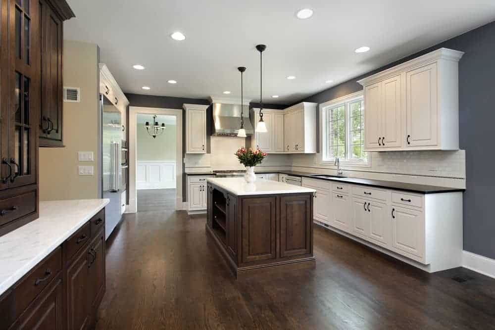 A spacious kitchen with white cabinets, a central island with dark wood cabinets, stainless steel appliances, and a vase with flowers. The room has recessed lighting, pendant lights over the island, and dark hardwood floors. Large windows let in natural light—a perfect example of Kitchen Design Orlando Florida.